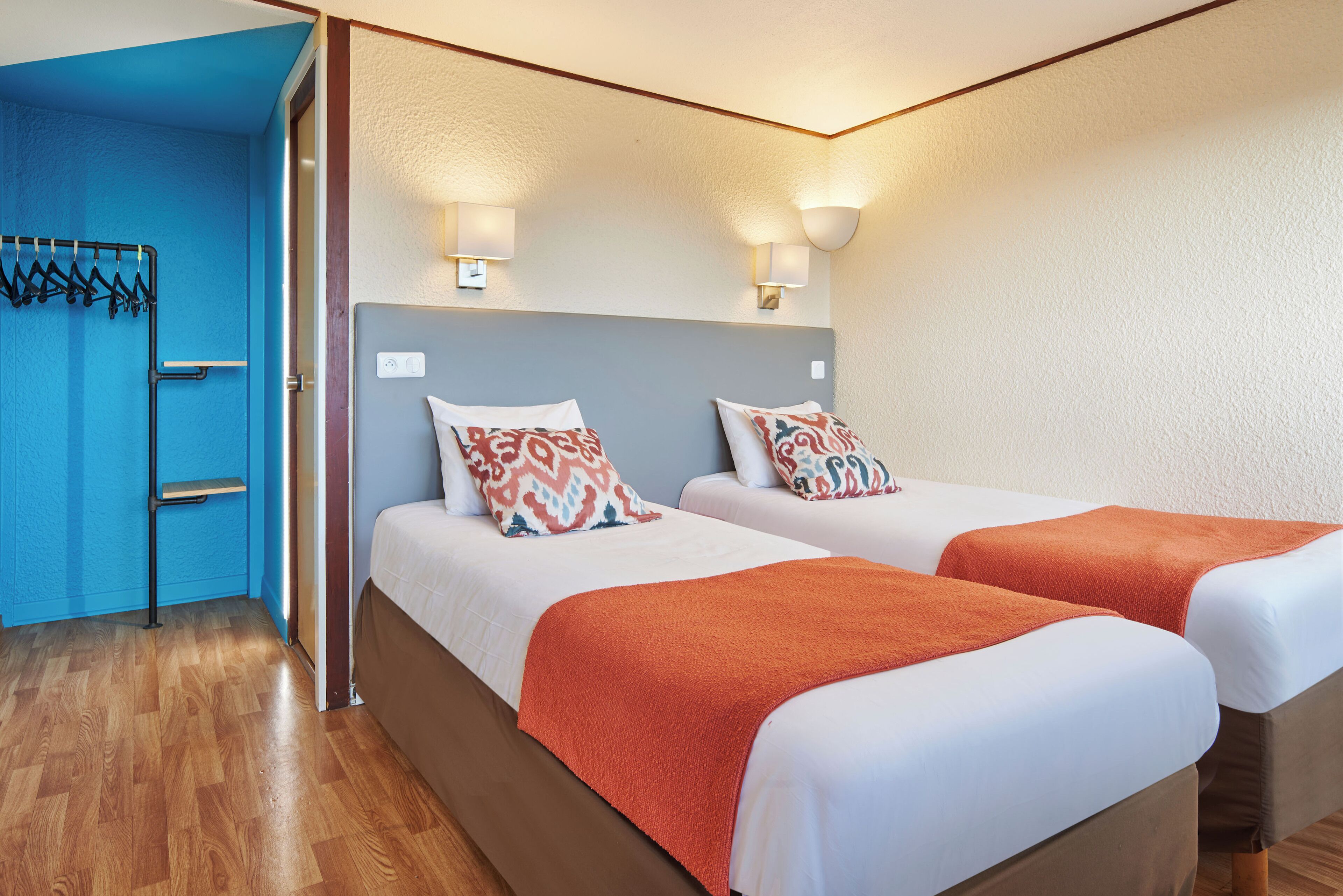 Ein Hotelzimmer mit zwei Betten, einer blauen Wand, Holzboden und einem Kleiderständer.