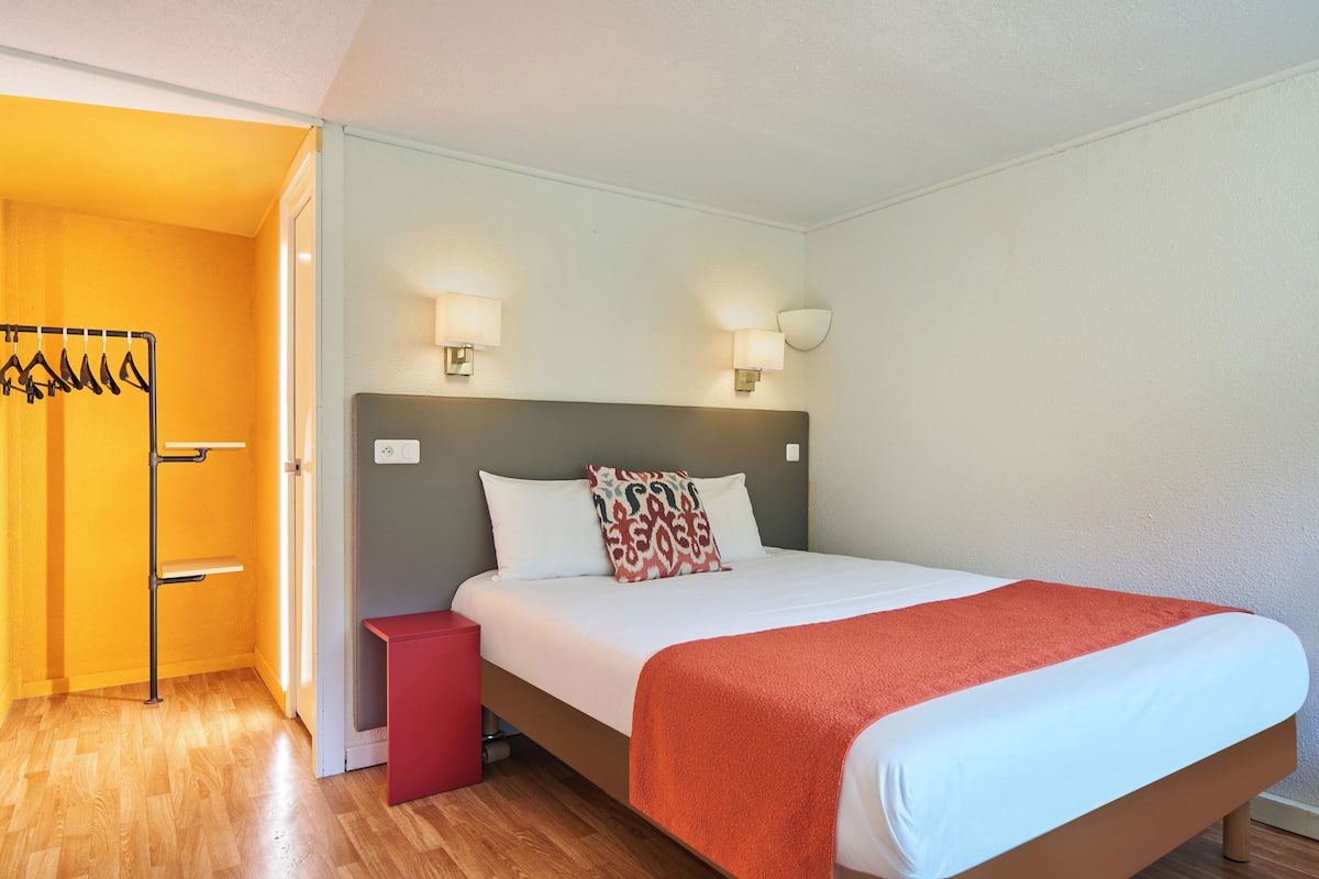 Quarto de hotel com uma cama, uma mesa de cabeceira vermelha, uma luminária na parede e um cabideiro.