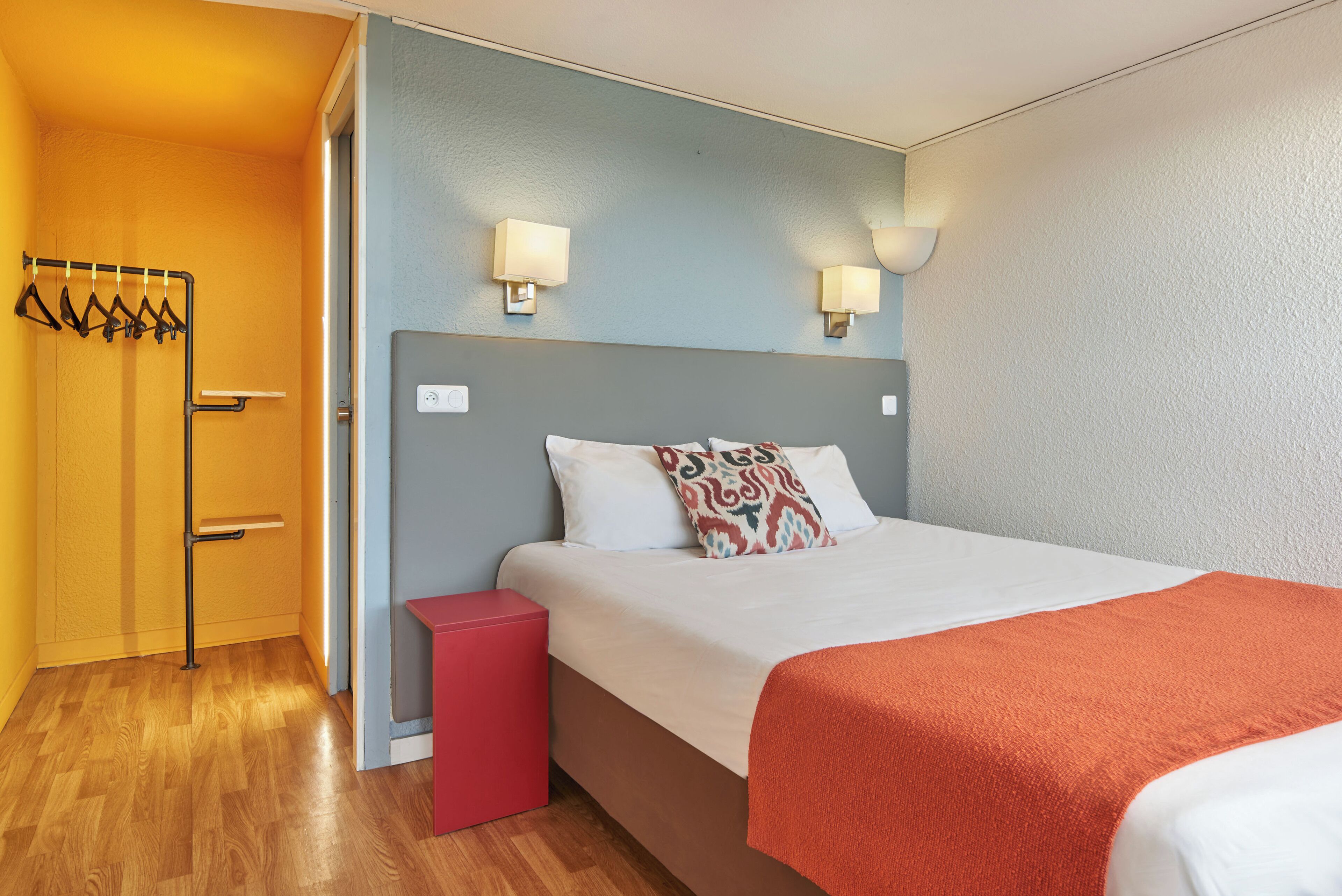 Camera d'albergo con un letto, un comodino rosso, una lampada a parete e un appendiabiti con grucce.