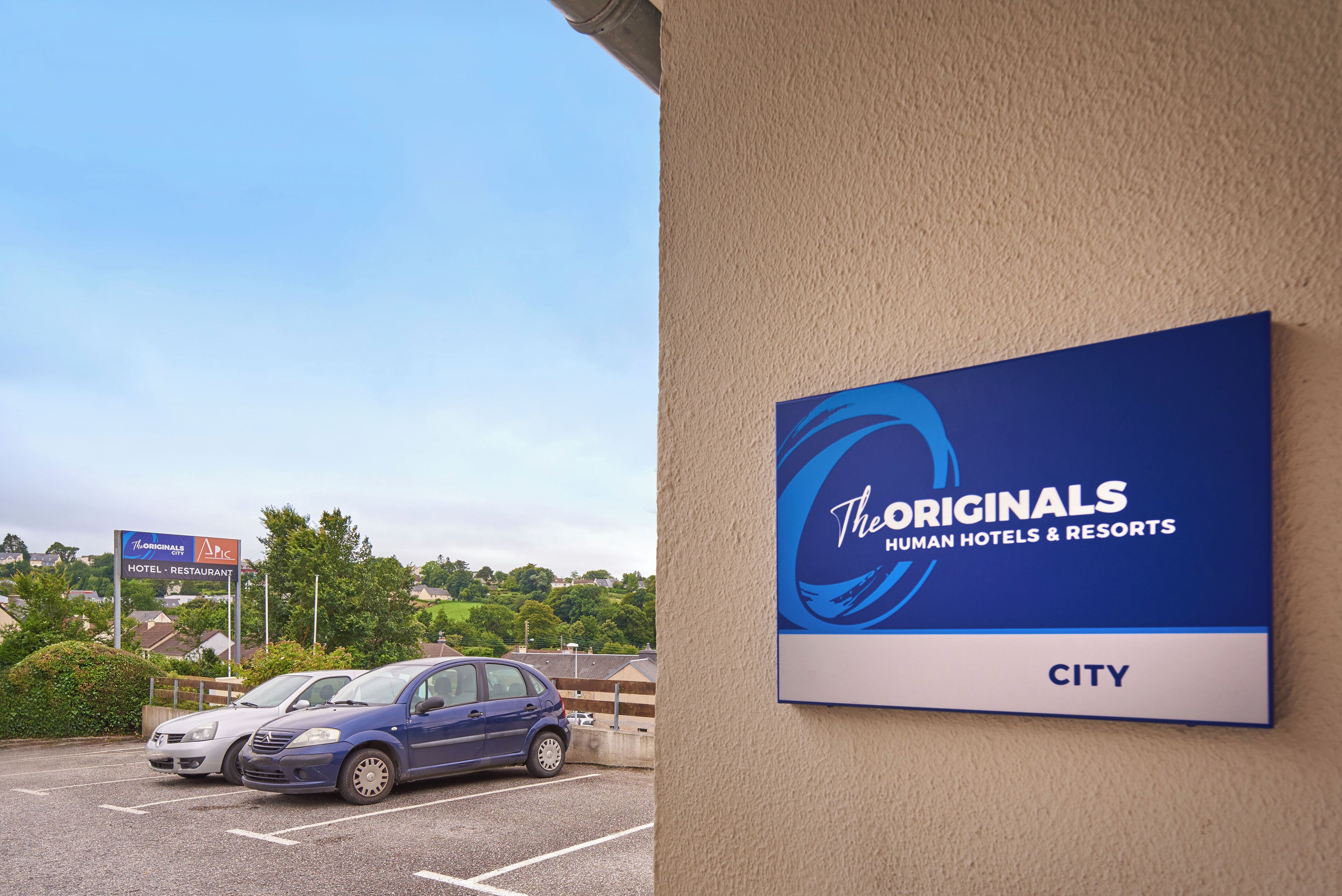 Une enseigne pour la chaîne d’hôtels The Originals Human Hotels & Resorts, avec un parking et une voiture au premier plan.