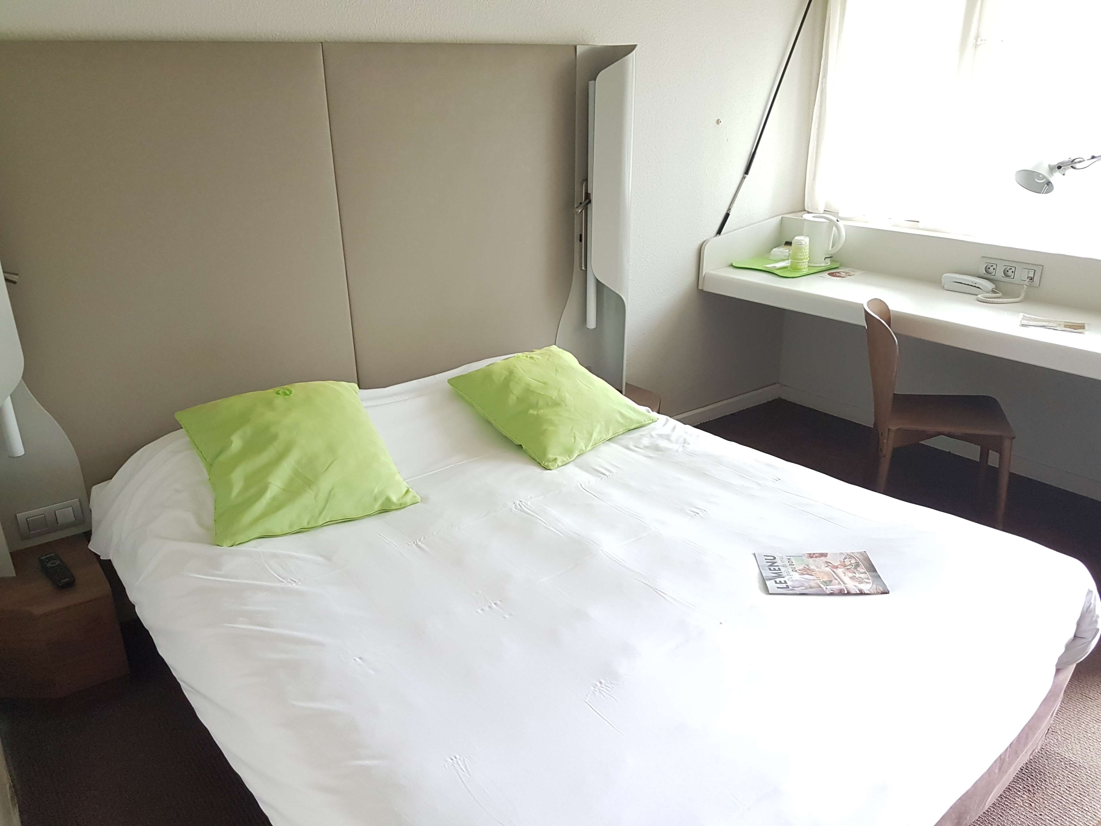 Ein Hotelzimmer mit einem Bett, zwei Kissen, einem Schreibtisch mit Stuhl, einer Lampe und einem Fenster.