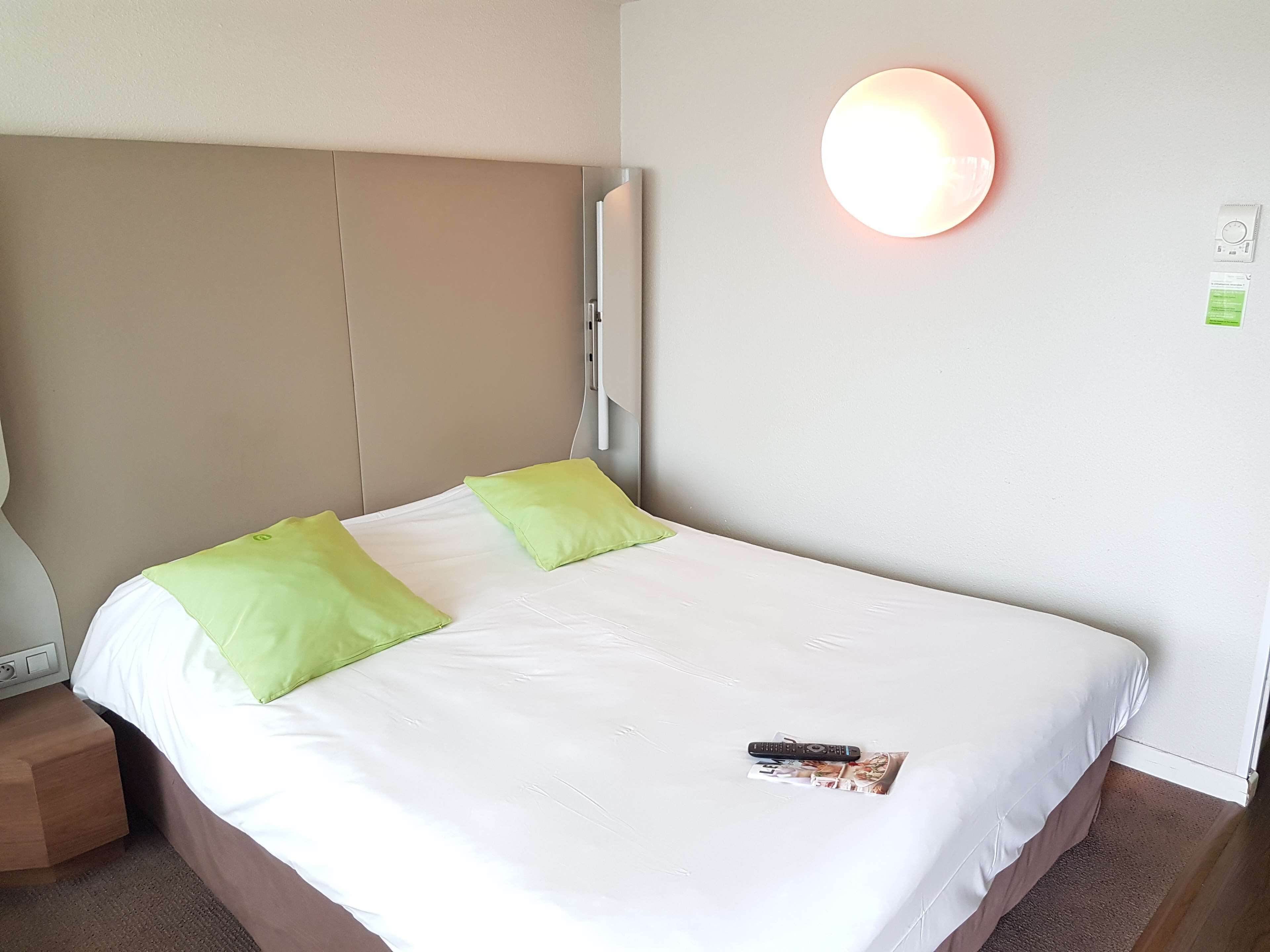 A hotel room with a large bed, two pillows, a remote control, and a magazine on the bed.