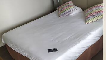 Ein Bett mit weißer Matratze, zwei gemusterten Kissen, einem Kopfteil und einem Nachttisch mit einem schwarzen Gegenstand darauf.