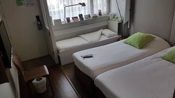A hotel room with two beds, a desk, a chair, a window with blinds, a lamp, and a small shelf with items.