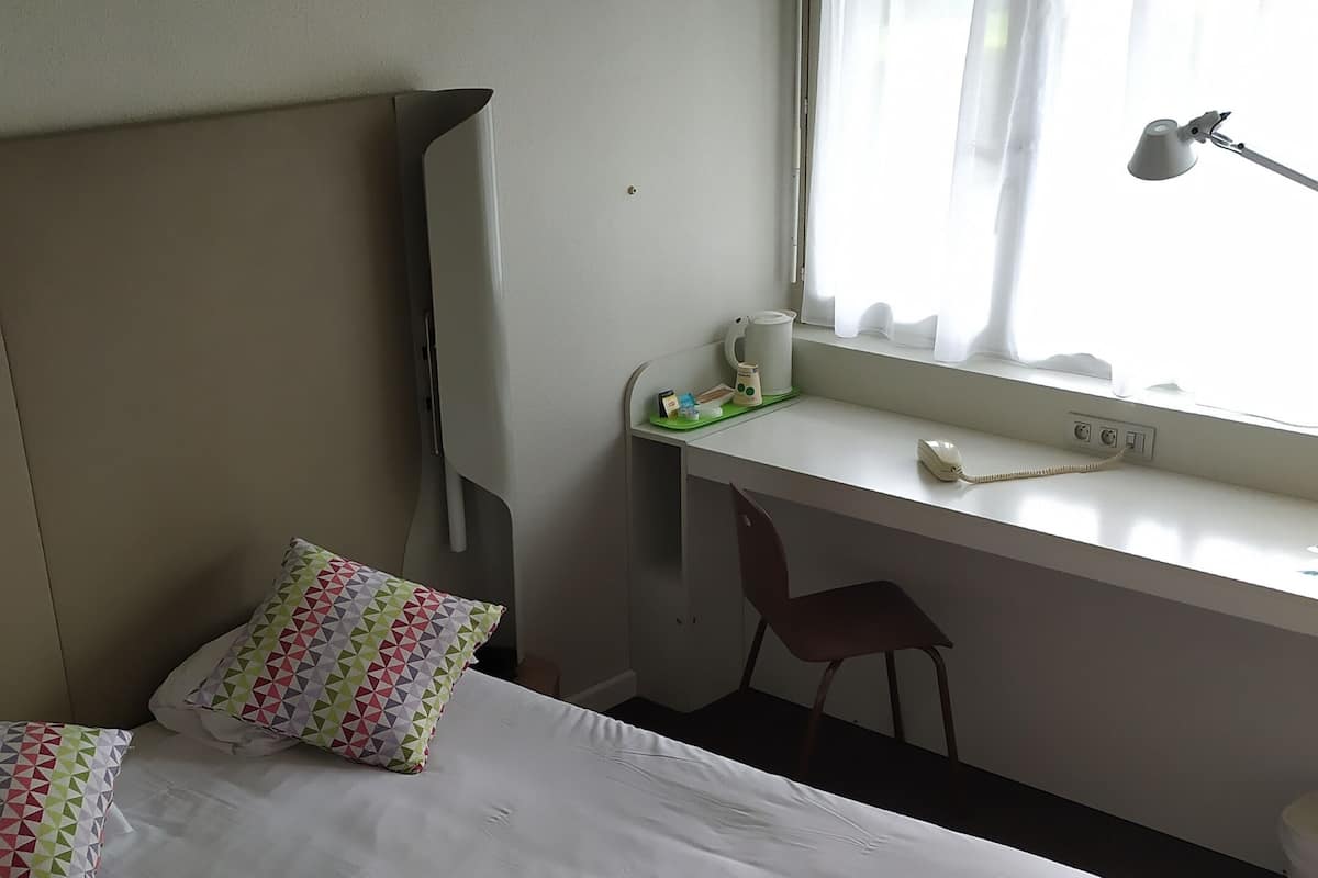 A neatly made bed with pillows, a desk with a lamp, and a chair in a room with a window.