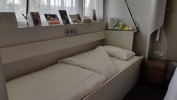 A single bed with a headboard, a bedside table with a lamp, a small shelf with a kettle and a phone, and a window with a view of trees and a building.
