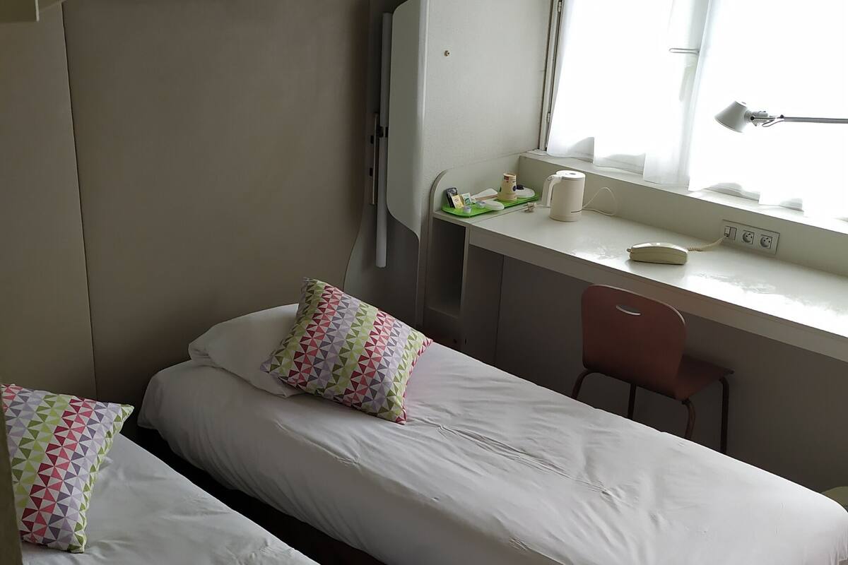 A compact hotel room with two single beds, a small desk with a chair, a window with curtains, and a ceiling fan.