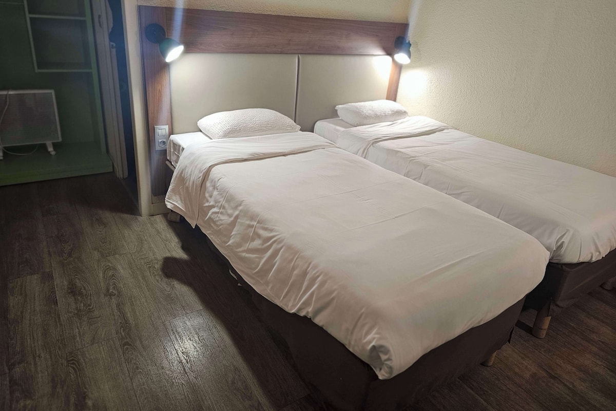 A room with two single beds, wooden flooring, a wooden headboard, and two wall-mounted reading lights.