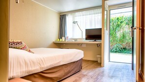 A bedroom with a bed, a desk with a TV, and a view of greenery through an open door. - Campanile Deauville St Arnoult (Saint-Arnoult)