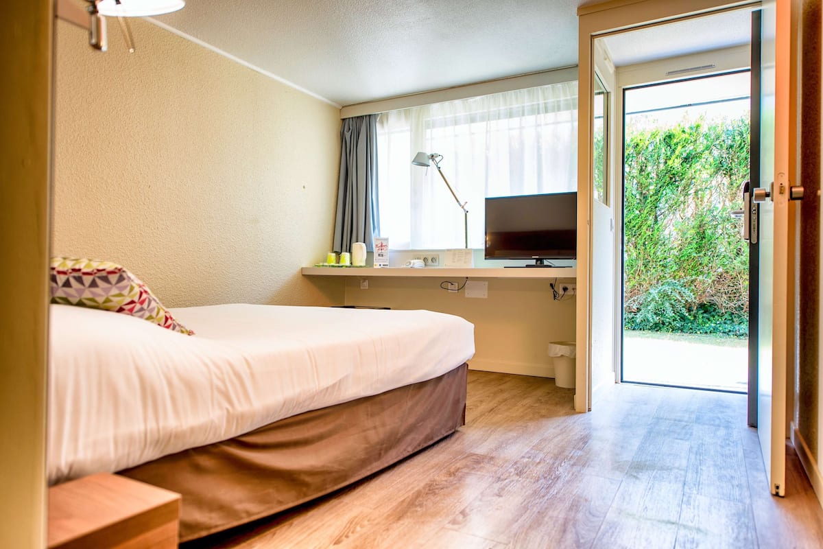 A bedroom with a bed, a desk with a TV, and a view of greenery through an open door.