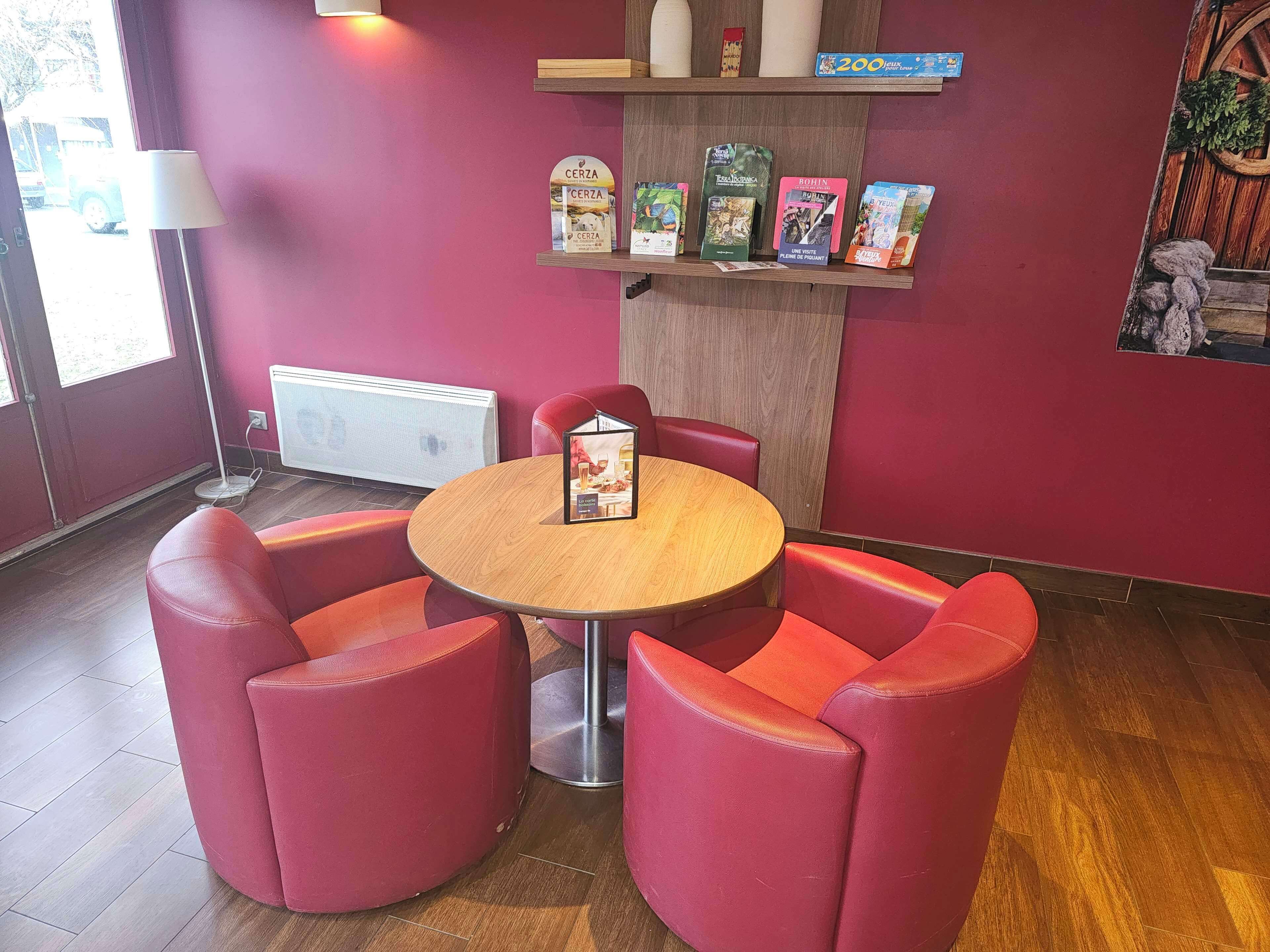 A cozy seating area with a round wooden table and red upholstered chairs. A small shelf above the table displays various books and a sign.