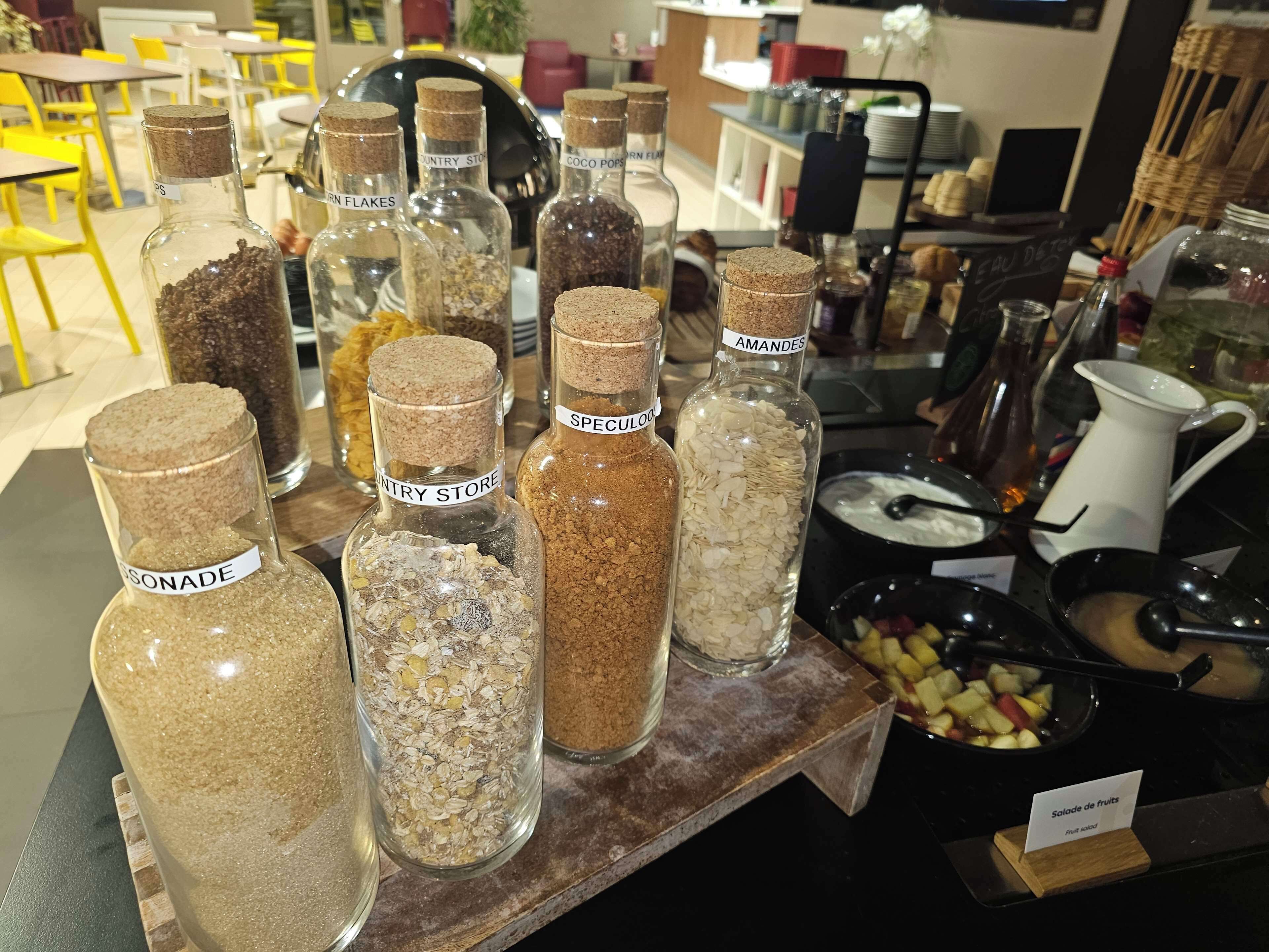 A display of various grains and seeds in glass jars with labels, a fruit salad sign, and a coffee pot on a table in a cafe setting.