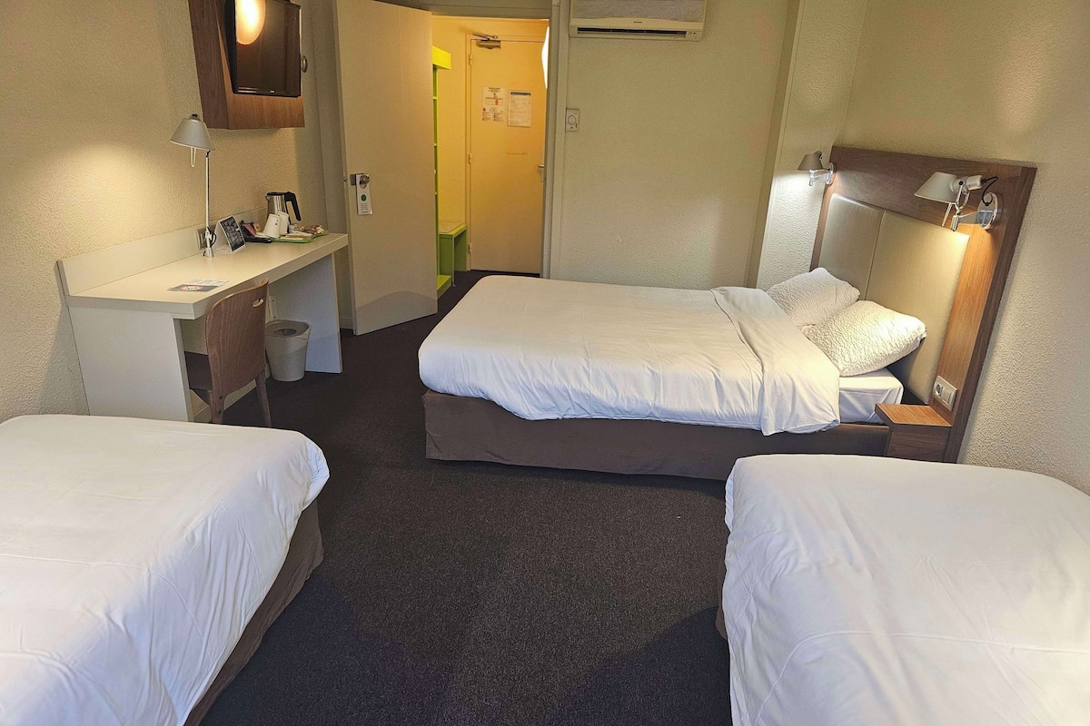 A hotel room with two single beds, a desk with a chair, a mirror, and a small bedside table with a lamp.