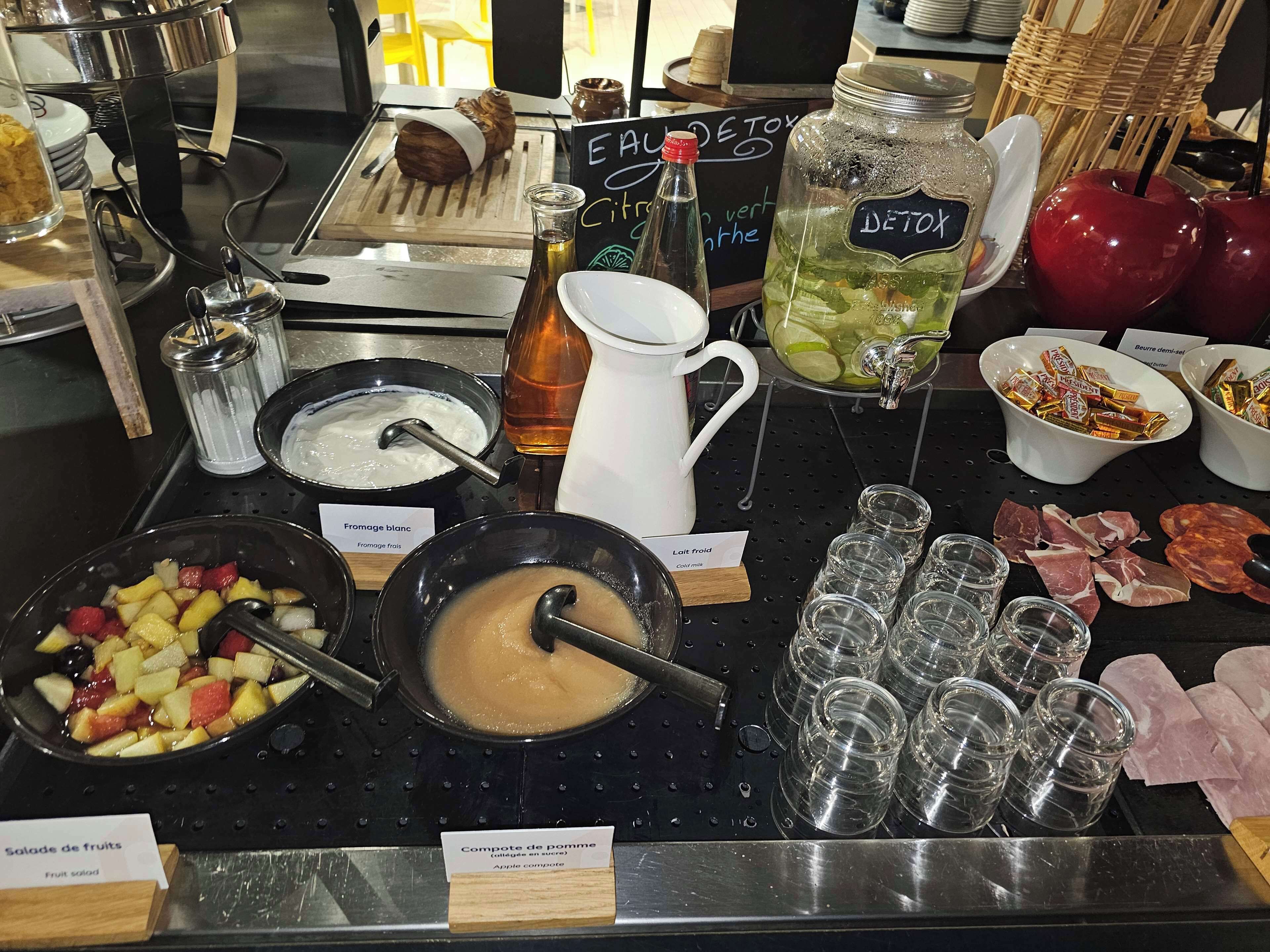 Buffet com diversos alimentos, incluindo uma salada de frutas, uma tigela de sopa e um pote rotulado 'DETOX'.