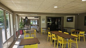 A spacious indoor dining area with multiple tables and yellow chairs, large windows on one side, and a kitchen area visible through an open doorway. - Campanile Deauville St Arnoult (Saint-Arnoult)
