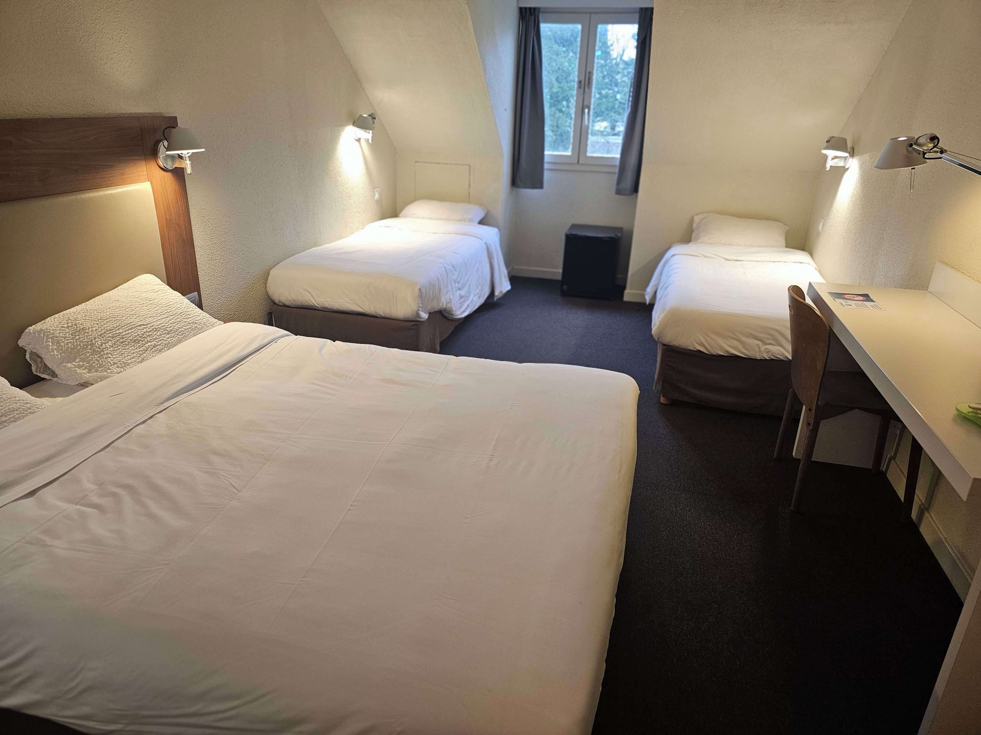 Habitación de hotel con dos camas, un escritorio y una silla. Se ve una ventana con cortinas y una puerta.