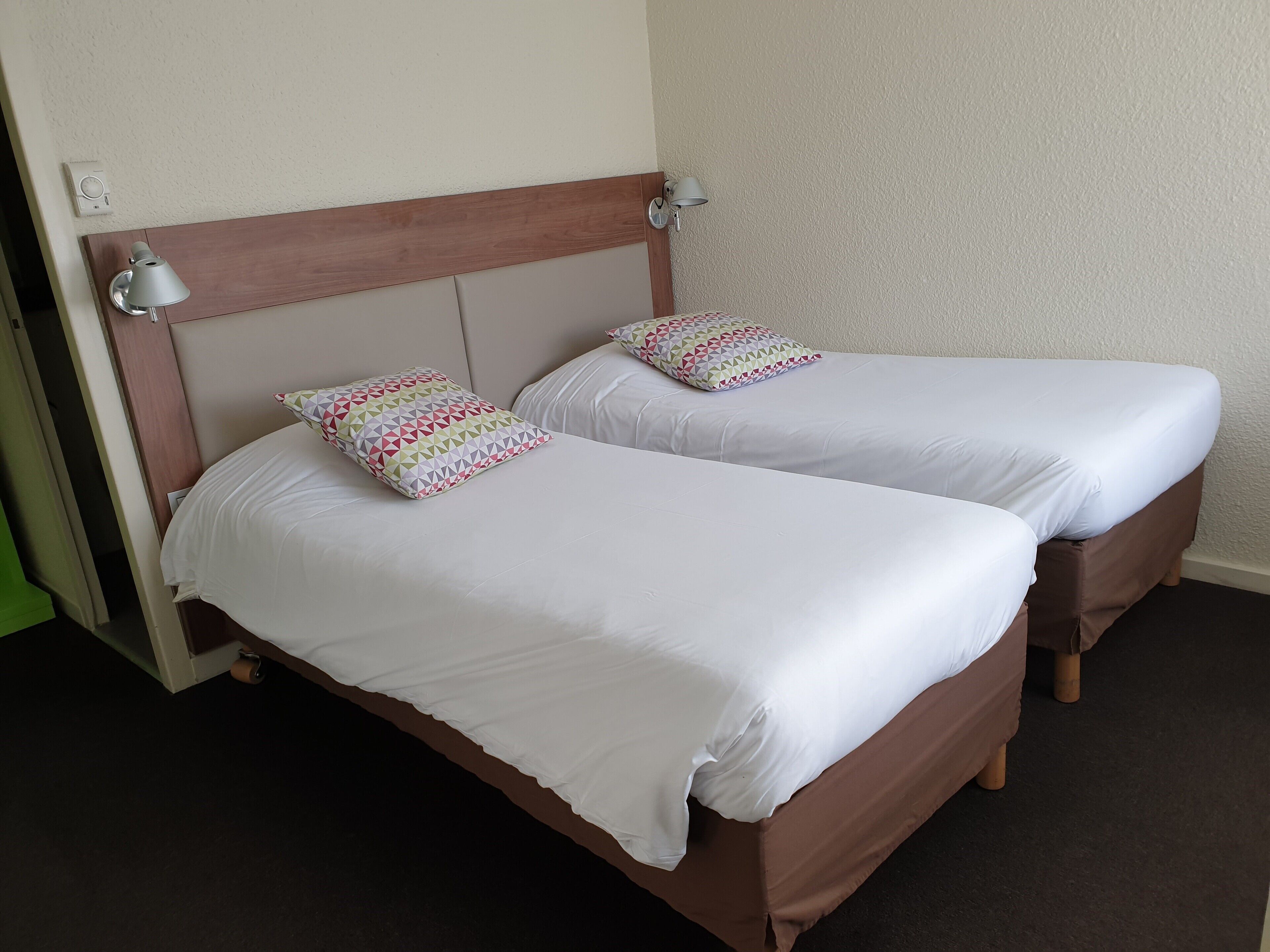 A double bed with a wooden headboard and two pillows, a bedside lamp, and a wall-mounted thermostat.