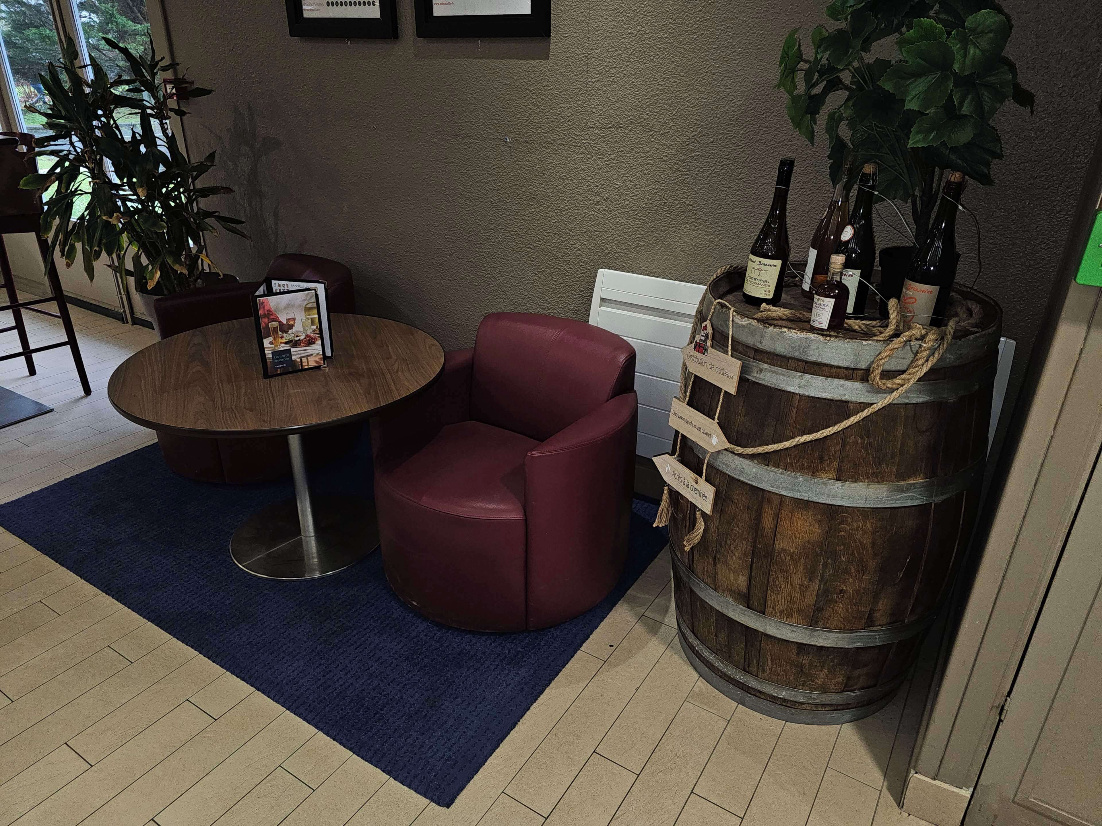 Een kamer met een ronde houten tafel, een rode stoel, een grote ton met flessen en een potplant.