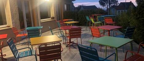 Un patio al aire libre con varias mesas y sillas, un edificio con techo inclinado y vistas del entorno.