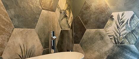 A bathroom with a white vessel sink, a wooden countertop, and hexagonal stone tiles with leaf patterns.