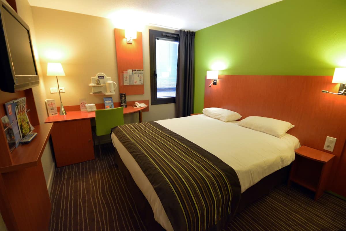A hotel room with a bed, bedside tables, a desk with a lamp, a television, and a window with curtains.