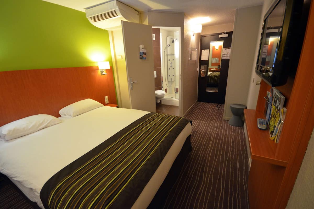 A hotel room with a bed, a bathroom with a shower, and a TV on the wall.