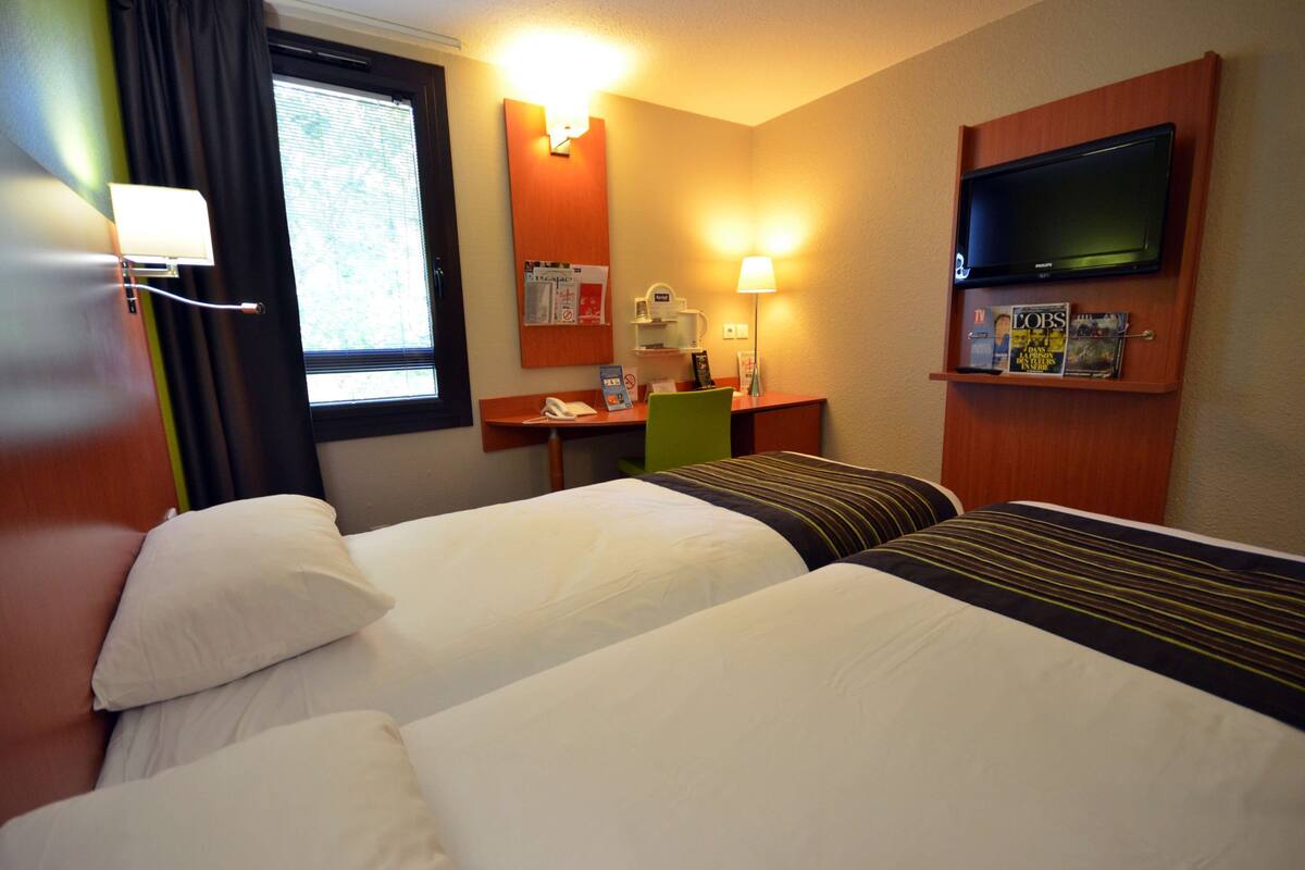 A hotel room with two beds, a desk with a telephone, a lamp, a TV mounted on the wall, and a bulletin board with notices.