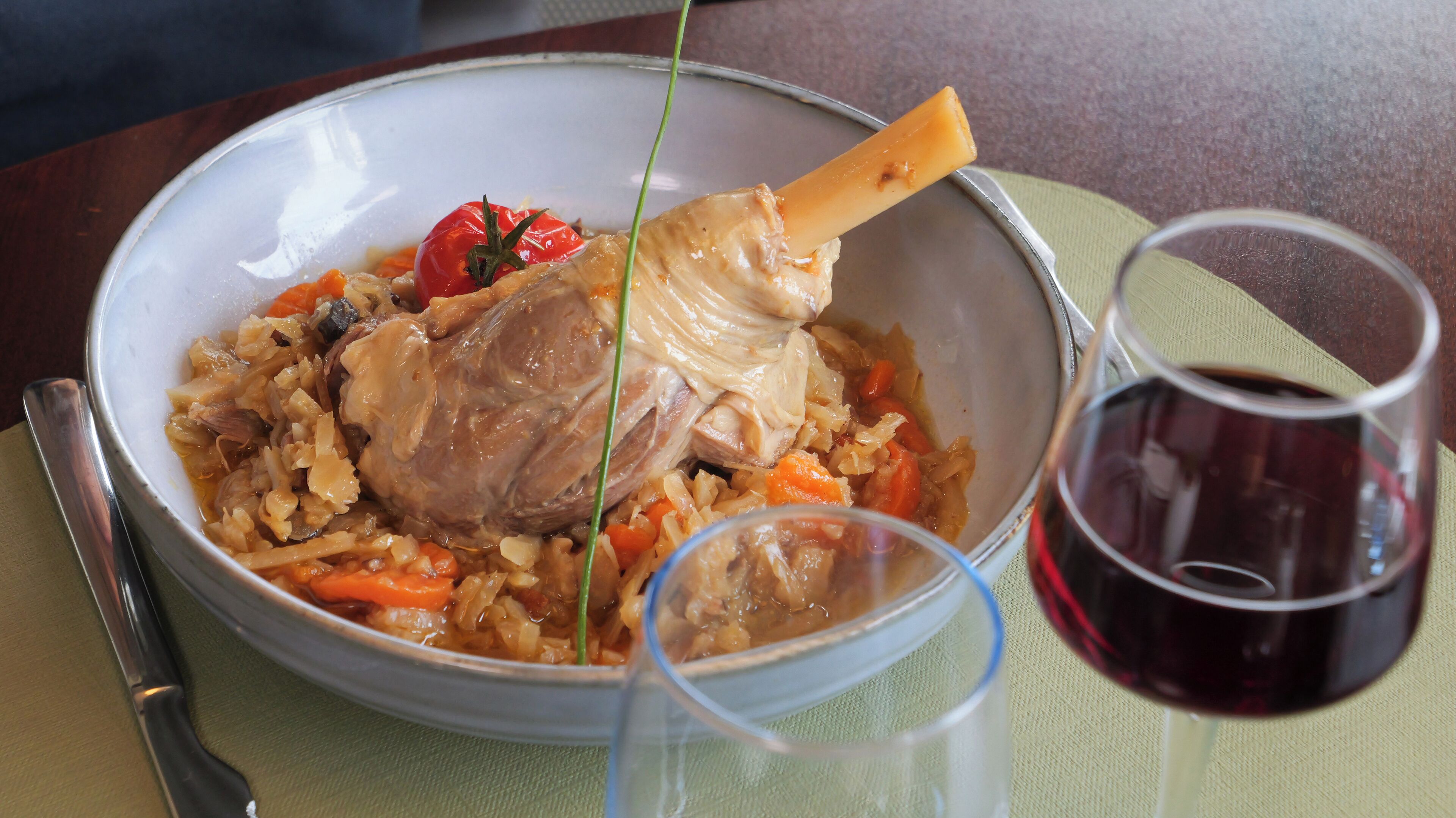 A bowl of food with a bone, vegetables, and a tomato on a plate. There are two glasses, one with a red liquid, possibly wine, and a spoon on the side.