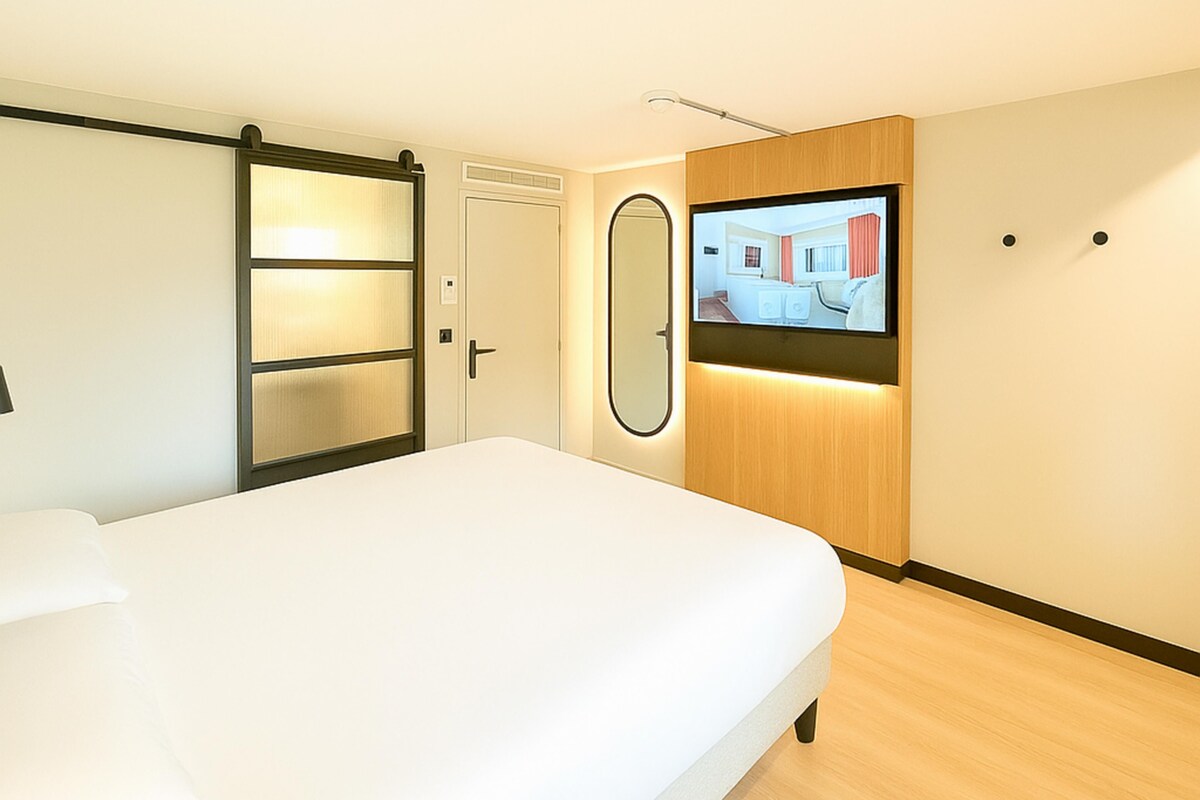 A hotel room with a bed, a wardrobe, a mirror, and a television screen displaying a room.