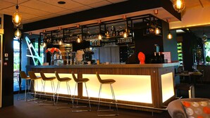 Zona de bar con mostrador de madera, taburetes, luces colgantes y estantes con botellas y vasos.