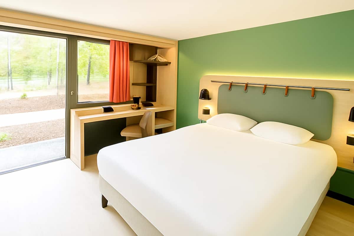 A modern hotel room with a large bed, a desk, and a sliding glass door to a balcony.