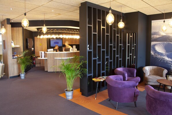 A reception area with a wooden reception desk, a television, and a large wall mural. There are potted plants and purple chairs. - Campanile NATURE - Clermont Ferrand Nord Riom (Riom)