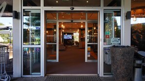 Una entrada de hotel con puertas de vidrio, un área de recepción con televisor y un espacio de estar con mesas y sillas.