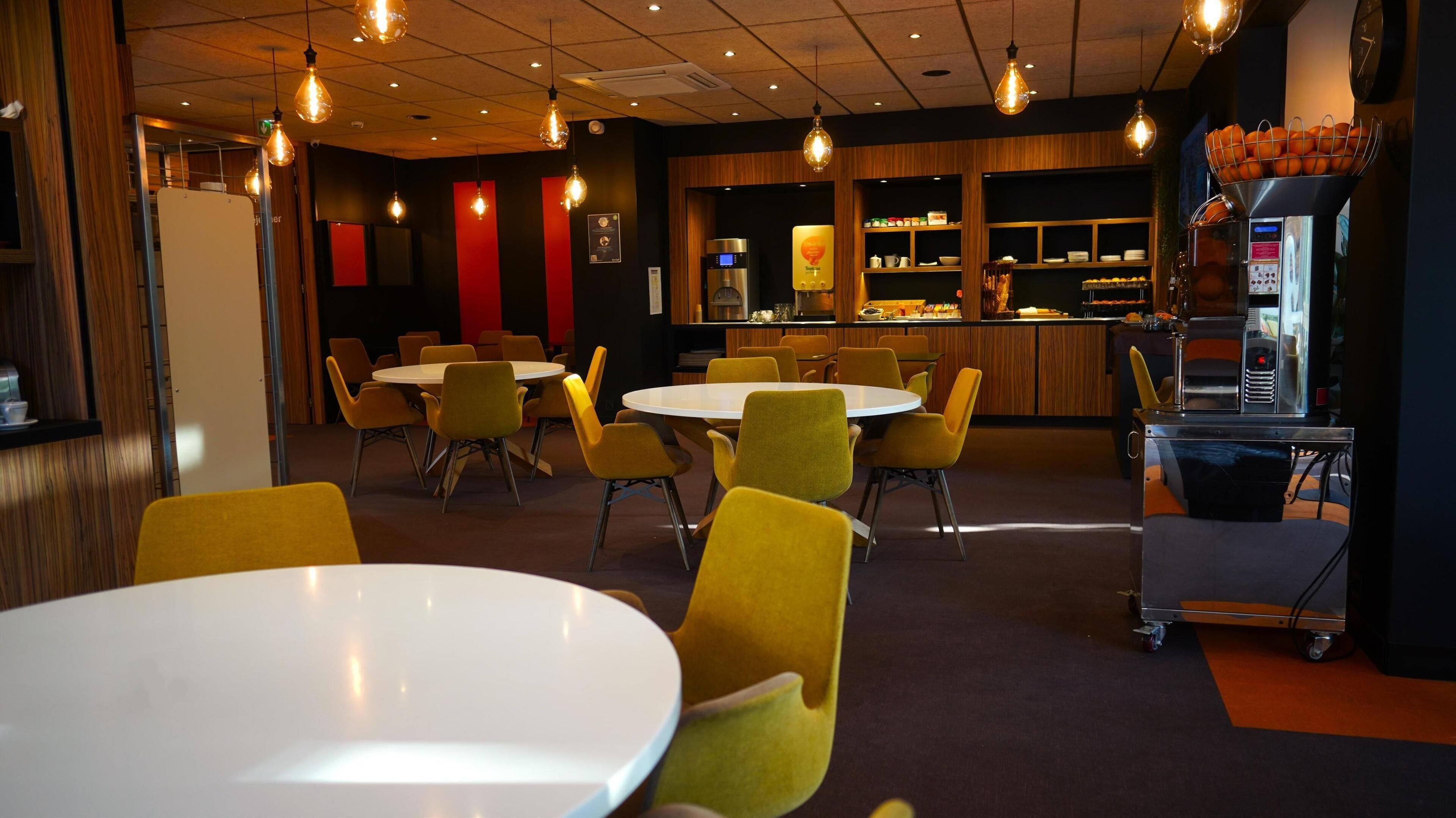 A restaurant interior with round tables and yellow chairs, a bar area with a coffee machine, and a fruit display.