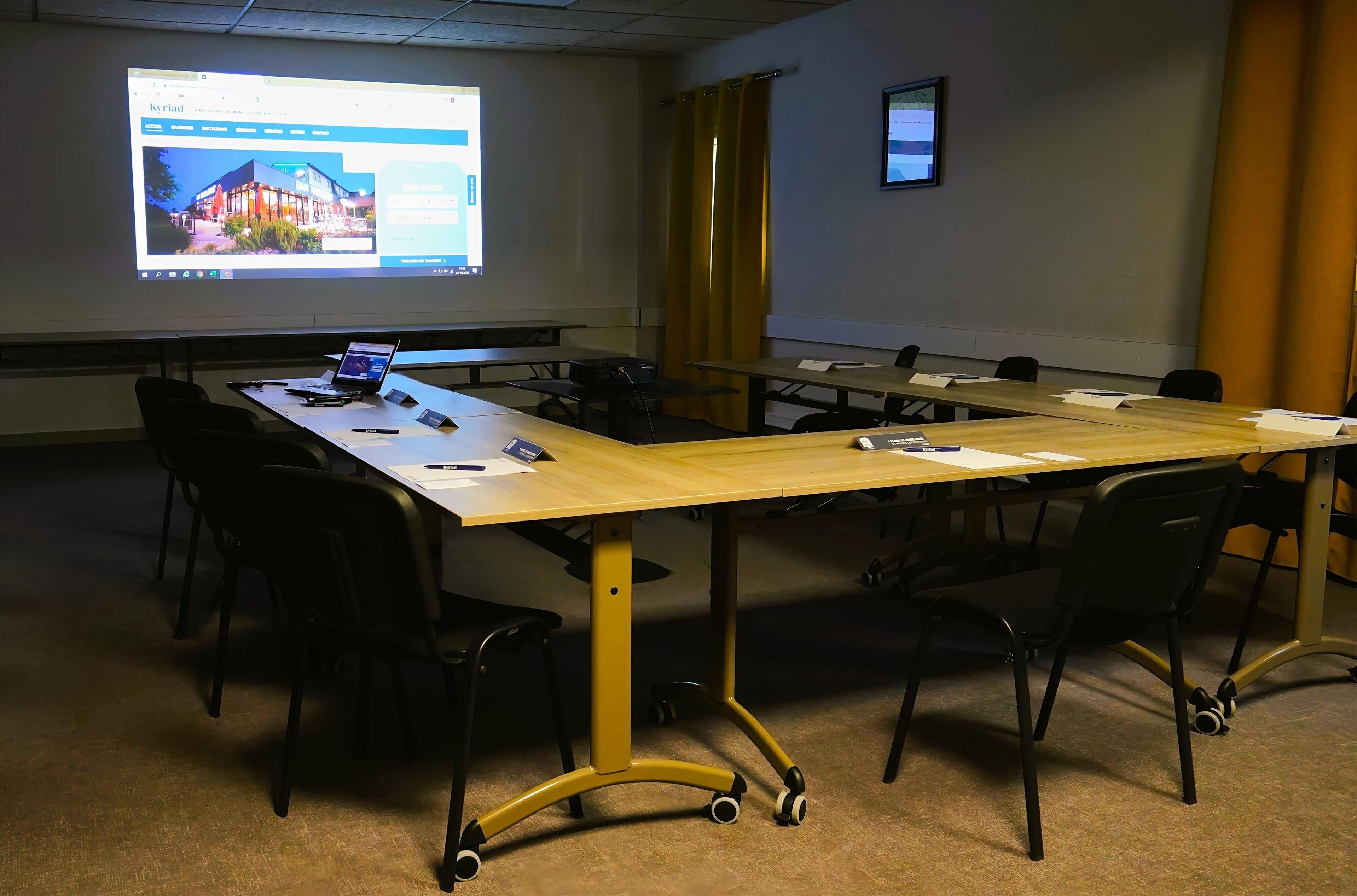 A conference room with a long table, black chairs, a laptop, papers, and a projector screen displaying a webpage.