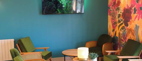 A room with a blue wall, a flat-screen TV, and a floral wallpaper. There are green chairs and a wooden table with a lamp.