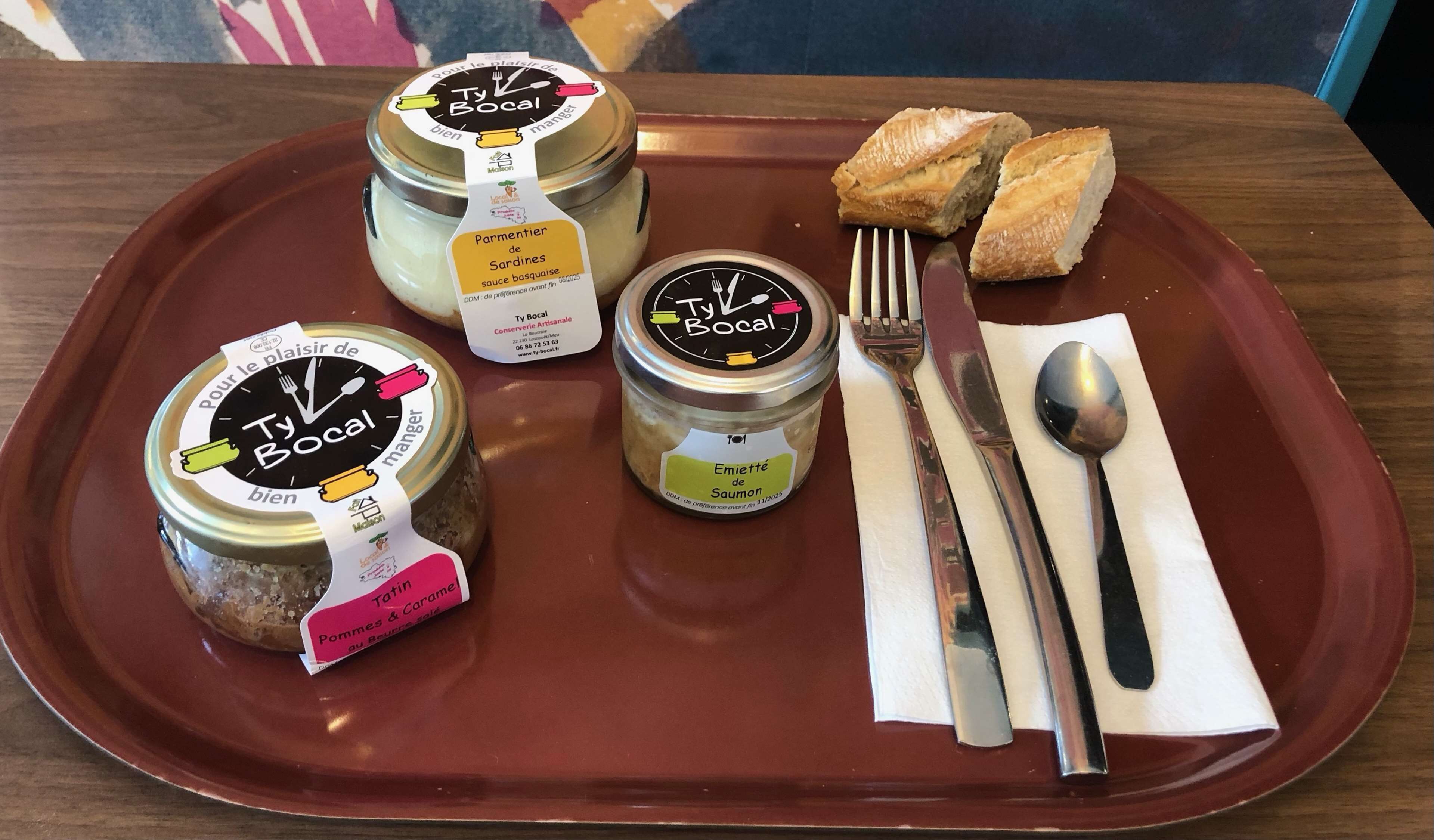 A tray with two jars of Ty Bocal preserves, a small dish of bread, and cutlery on a wooden table.