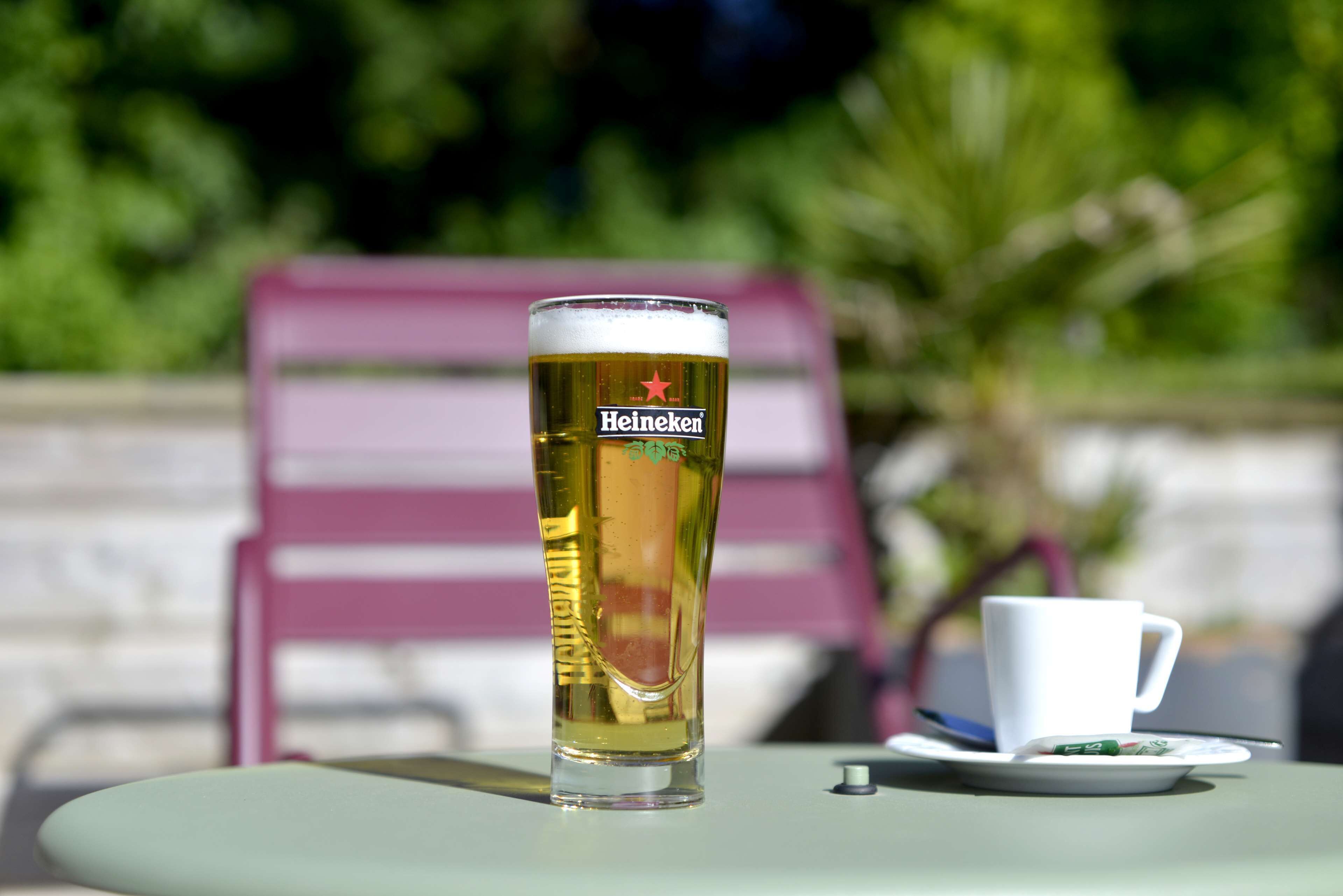 A Heineken glass on a table with a white cup and saucer beside it.