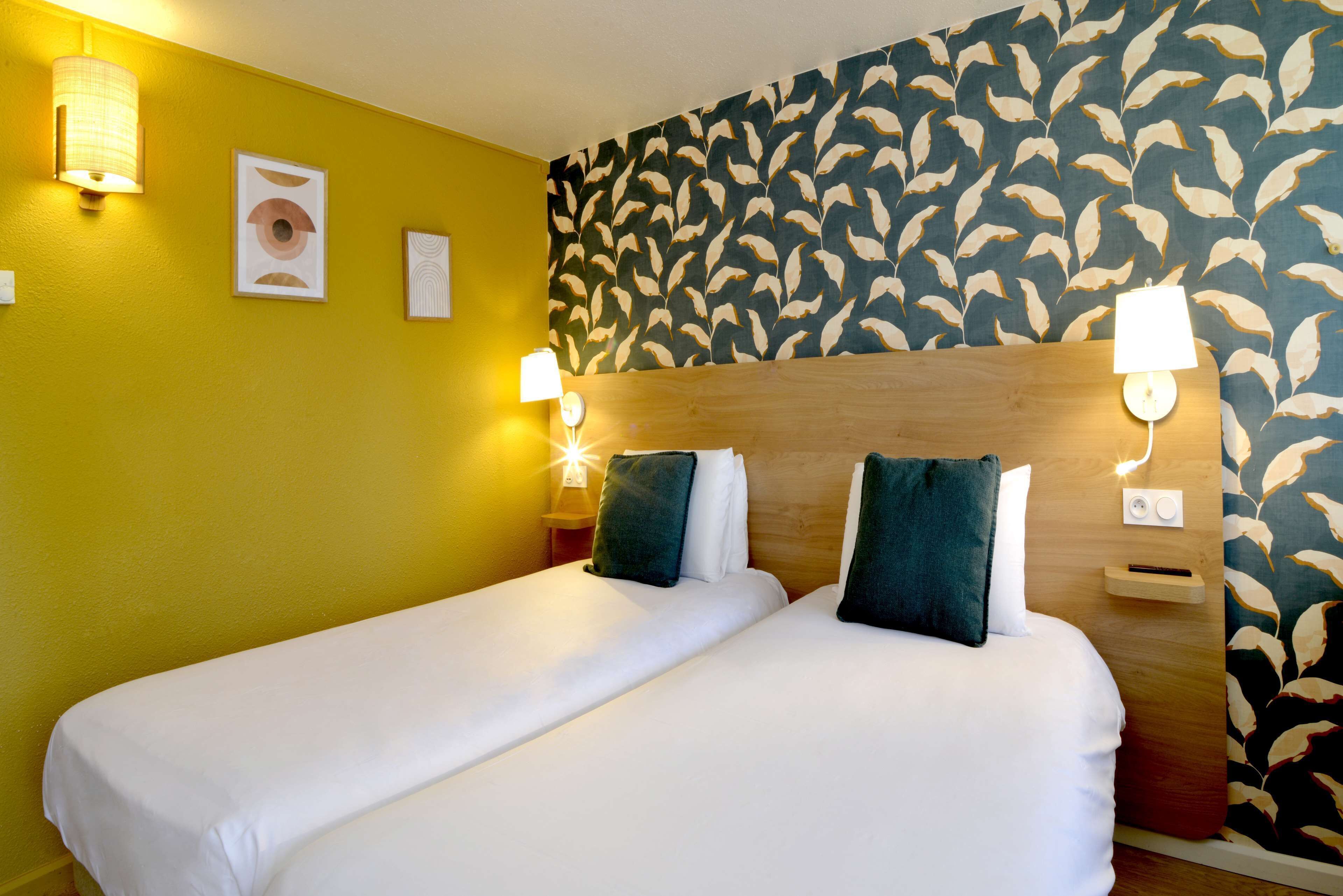 Una cama doble con sábanas blancas, dos mesitas de noche con lámparas, un cabecero de madera y una pared con un diseño de hojas.