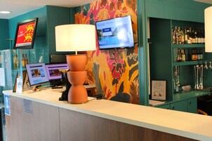 Un mostrador de recepción con una computadora, una lámpara y un televisor que muestra una contraseña de Wi-Fi.