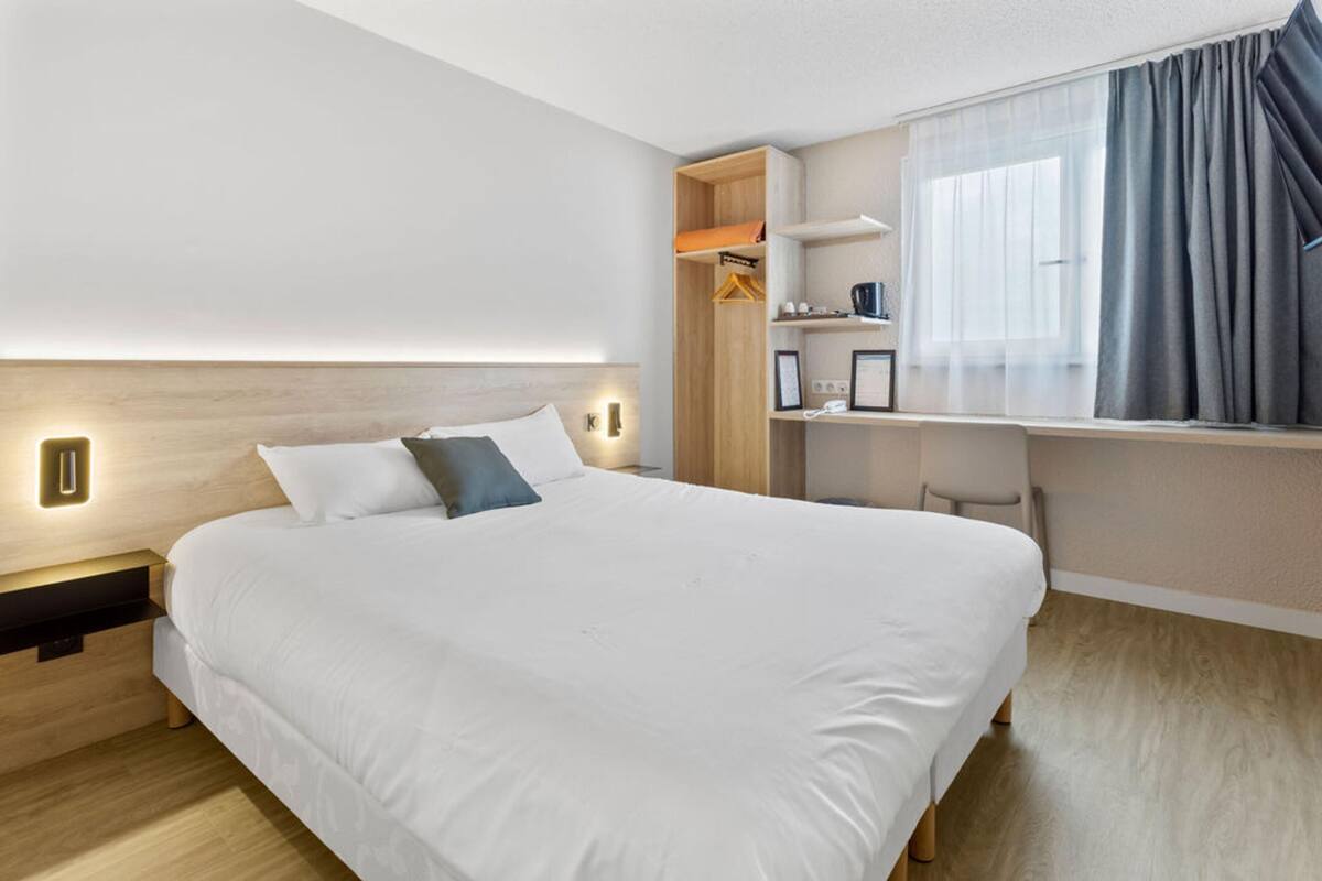 A modern hotel room with a large bed, bedside tables, a desk with a chair, and a window with curtains.