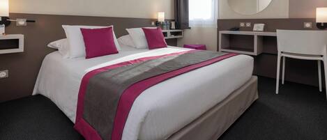 A hotel room with a large bed, two pillows, a desk, a chair, and a small table with a lamp.