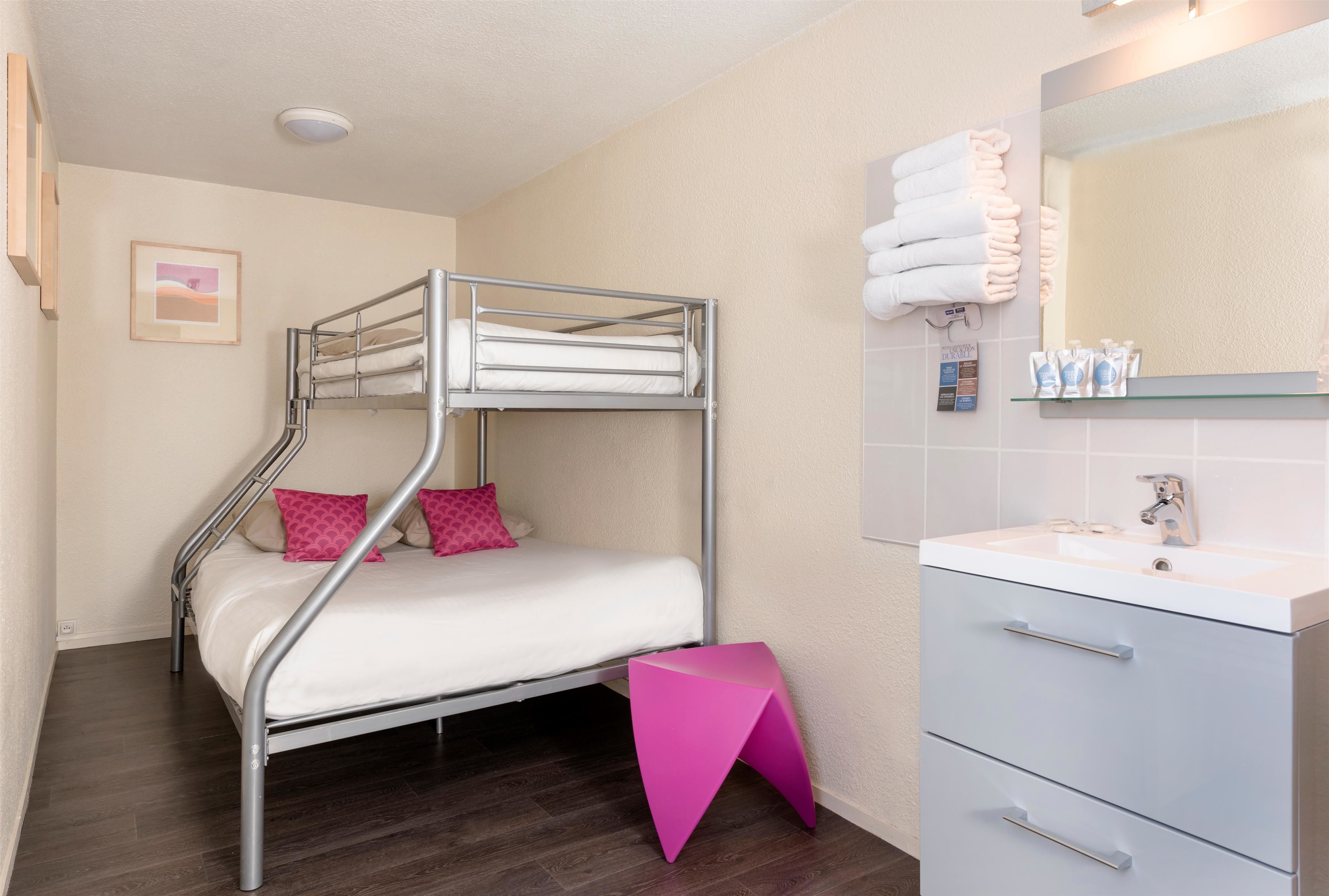A bunk bed with pink pillows, a small pink stool, a white sink, and a mirror with towels.