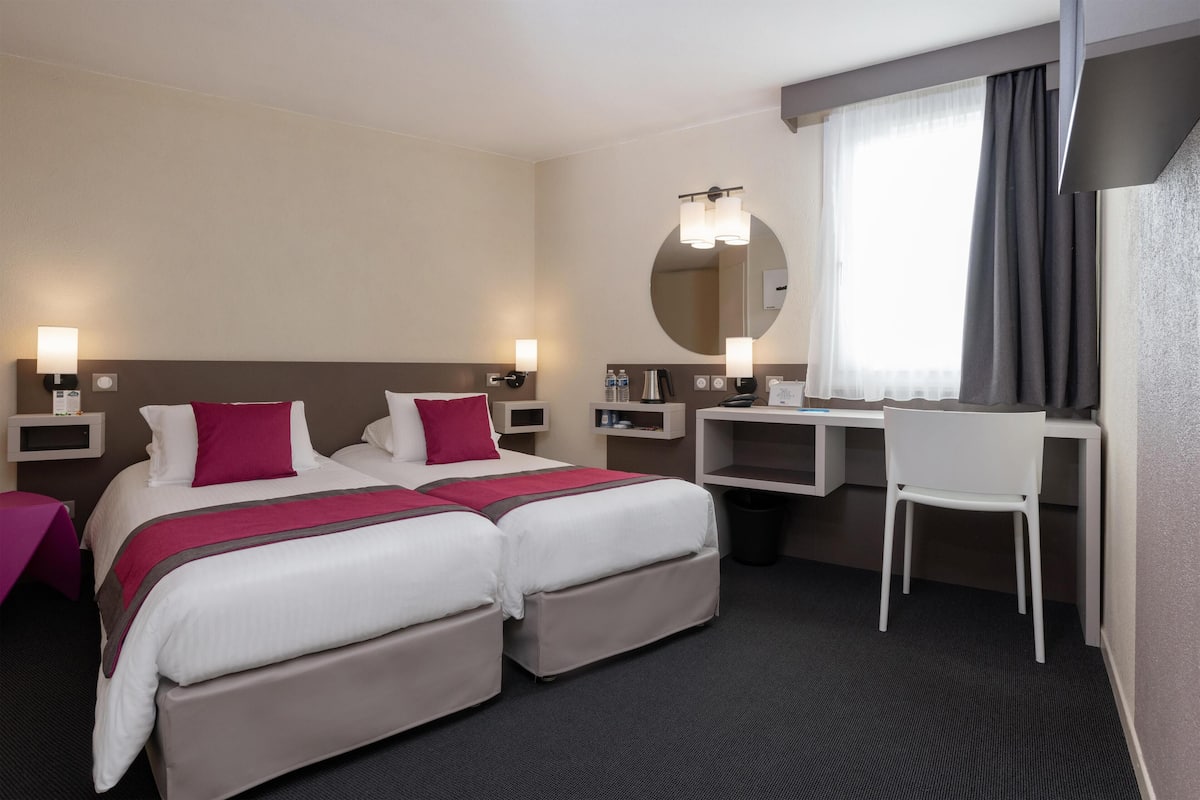 A hotel room with two beds, a desk, a chair, a mirror, and a window with curtains.