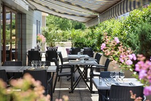 Un área de comedor al aire libre con mesas y sillas, rodeada de follaje y flores.
