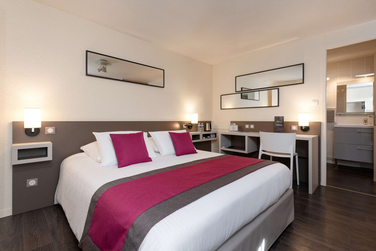 Habitación de hotel con una cama grande, un escritorio y una silla. Hay luces fijadas a la pared y un espejo sobre el escritorio.
