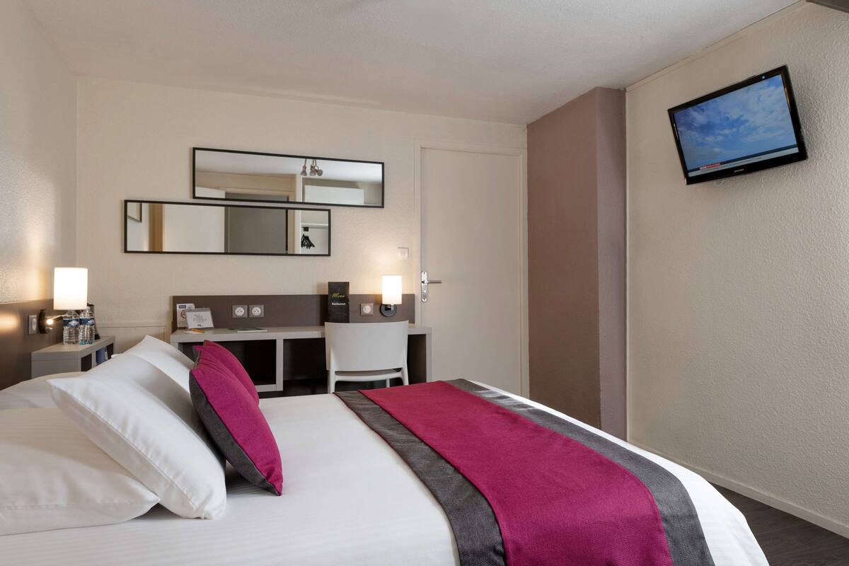 A hotel room with a bed, a desk, a chair, a TV mounted on the wall, and a mirror above the desk.