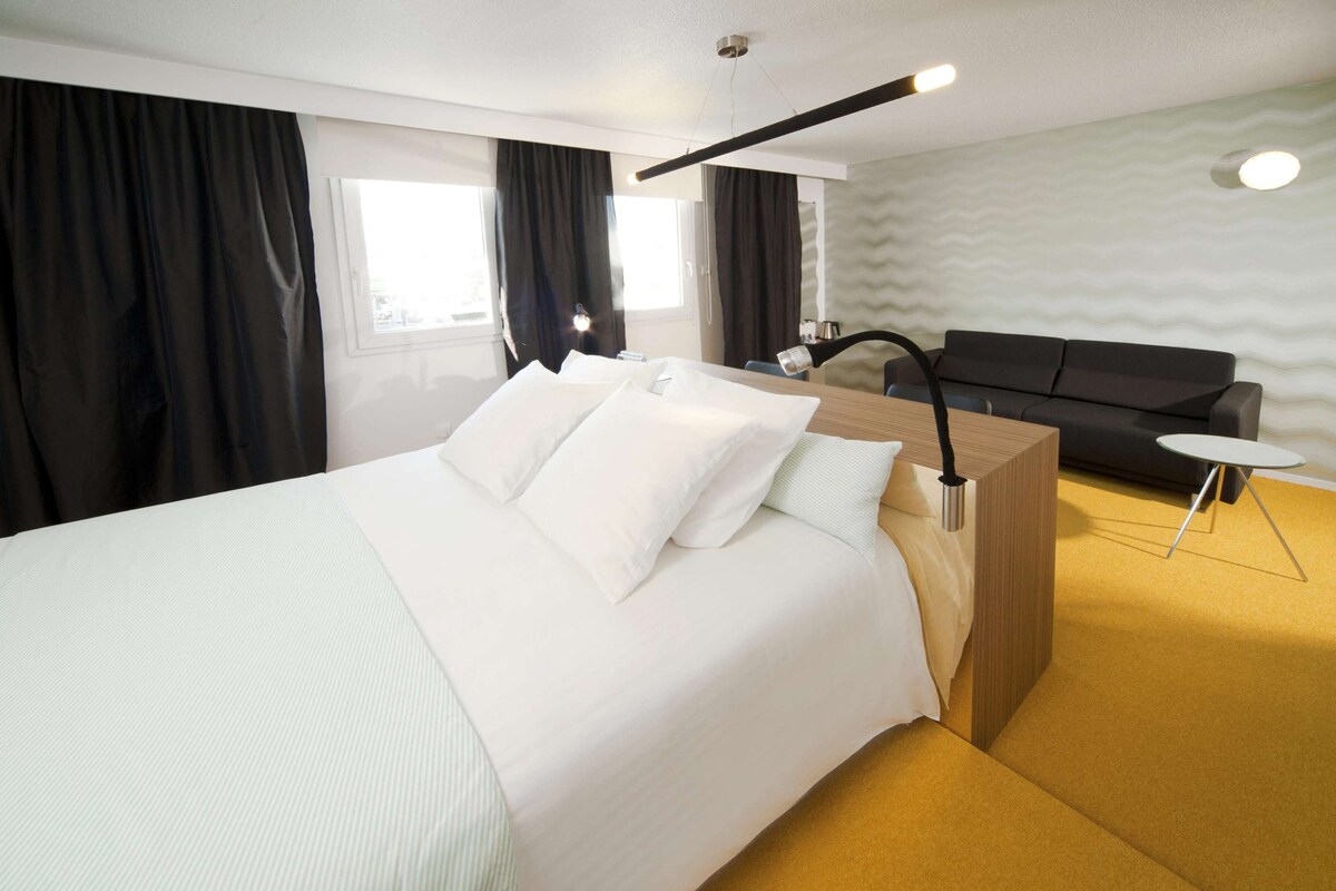 Una habitación de hotel moderna con una cama grande, una mesita de noche de madera, una pequeña mesa redonda, un sofá y una pared con un estampado ondulado.