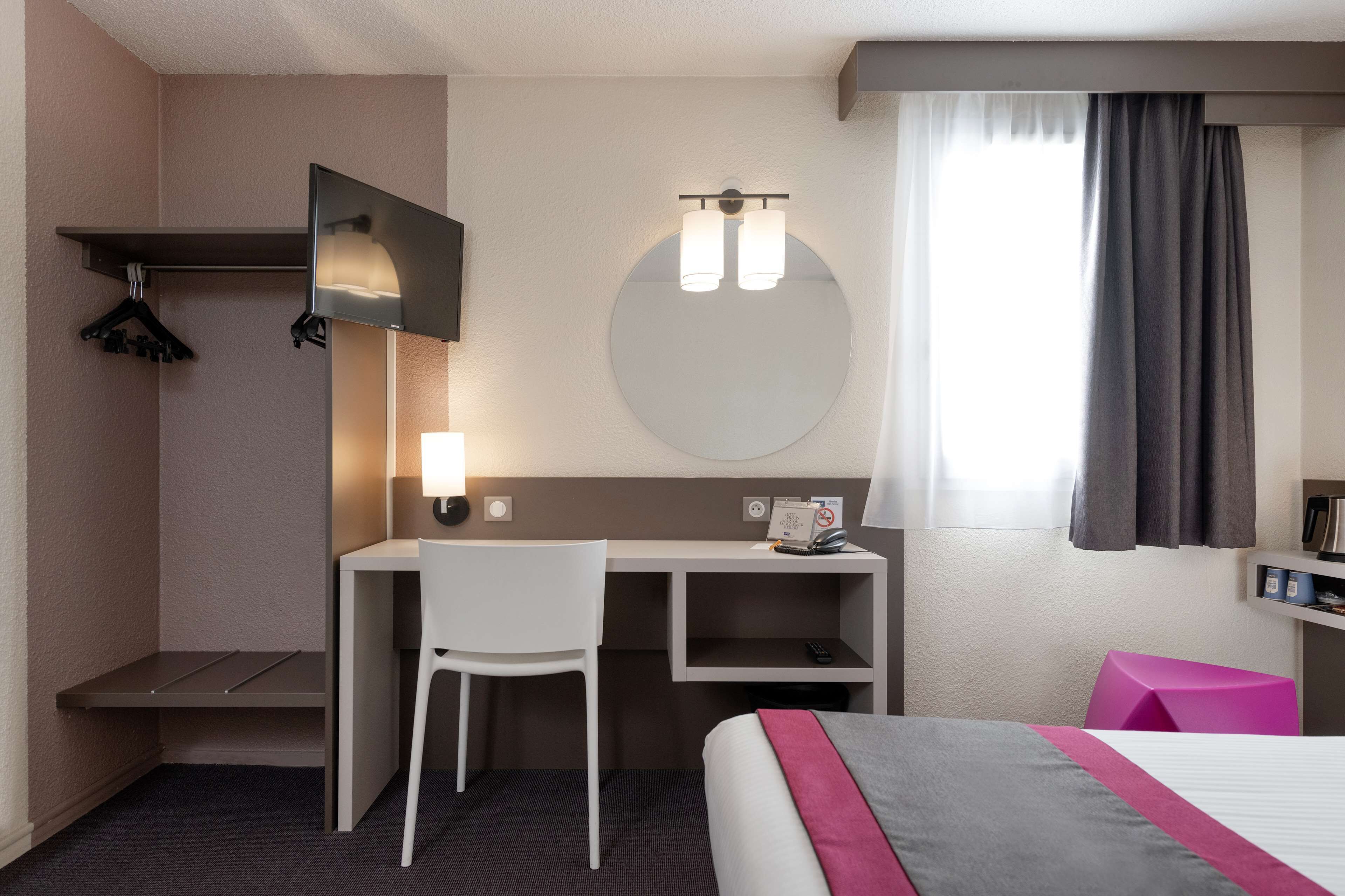 Habitación de hotel con cama, escritorio, silla, espejo, reloj de pared redondo y ventana con cortinas.
