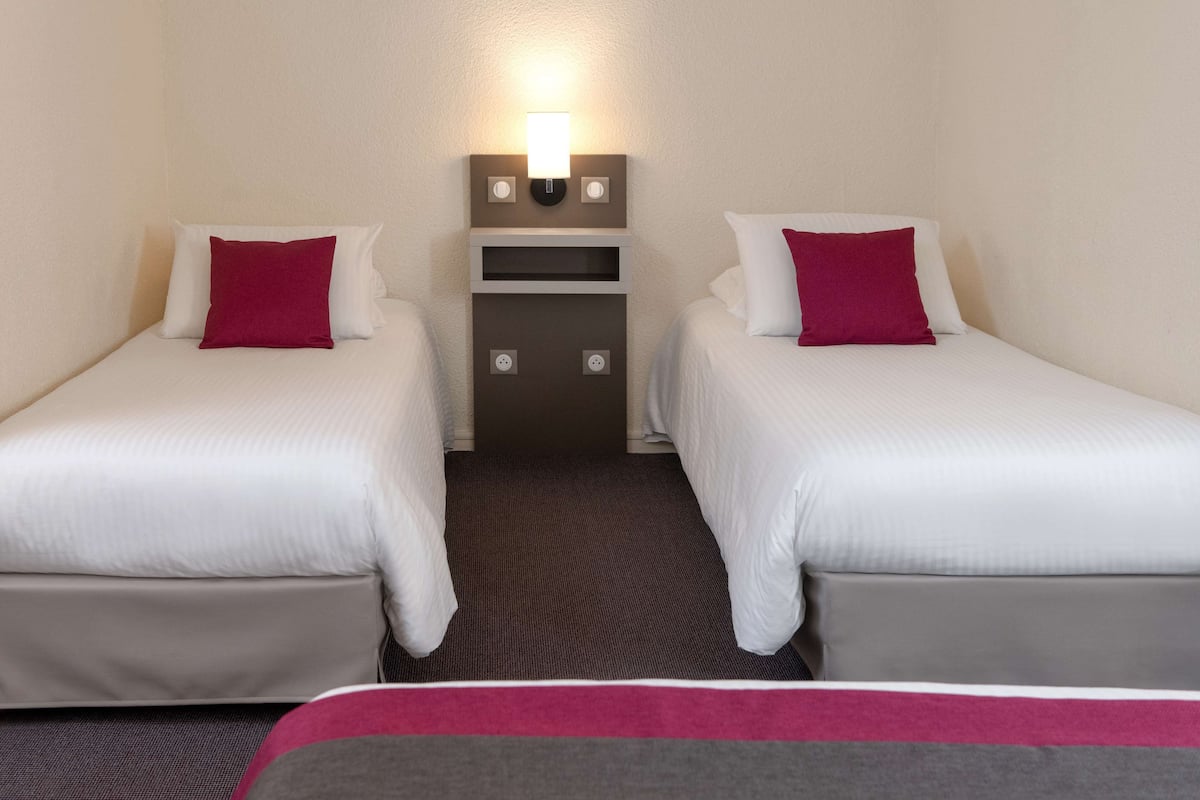 A hotel room with two single beds, each with a white mattress and a red pillow. There is a small nightstand with a lamp between the beds.